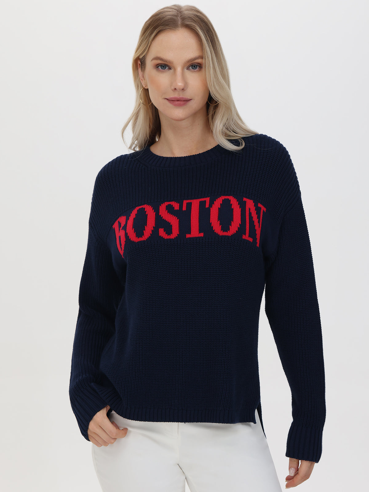 A person with long blonde hair wears a navy 525 America Boston sweater with a relaxed fit and red chest lettering. They pair it with white pants, gazing forward neutrally against a plain white background.