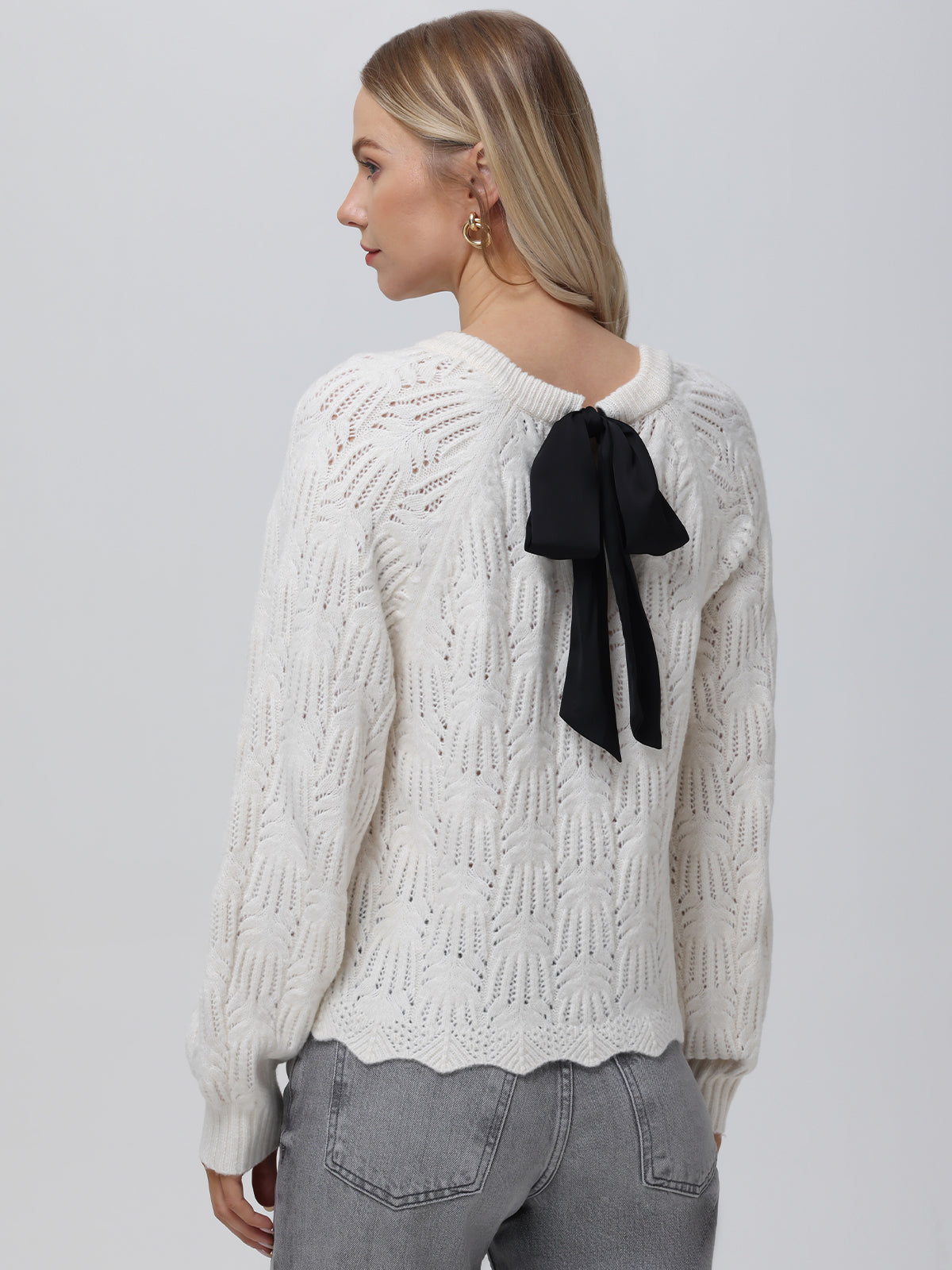 A blonde woman stands against a light background wearing a white 525 America knit pullover. The sweater features a black bow at the back, subtle pointelle details, and a crew neckline, stylishly complemented by her gray jeans.