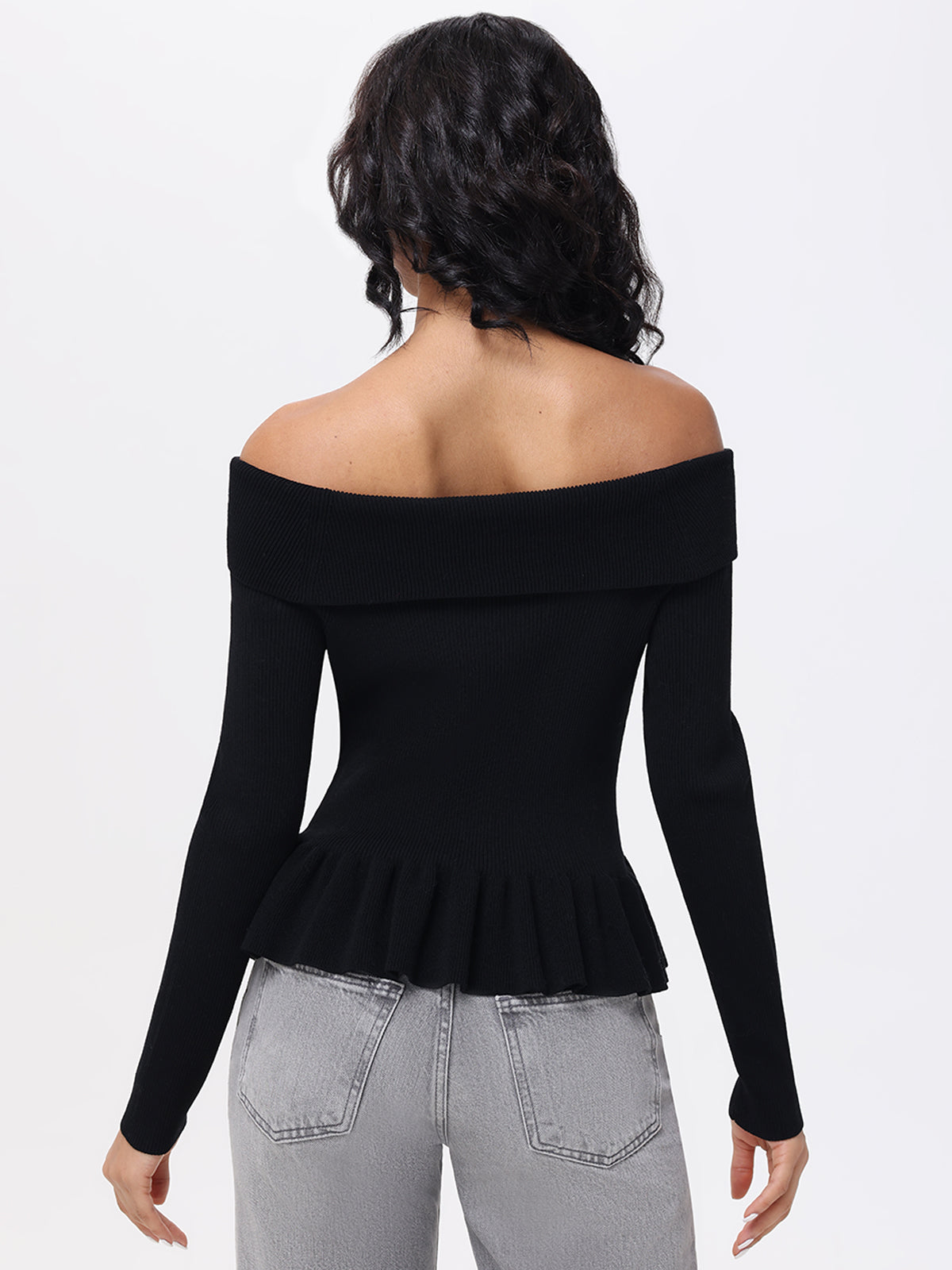 A person with long curly hair, facing away from the camera, is wearing an elegant 525 America Off Shoulder Peplum Pullover. The chic black top has long sleeves and a pleated hem and is paired stylishly with light gray jeans against a plain white background.