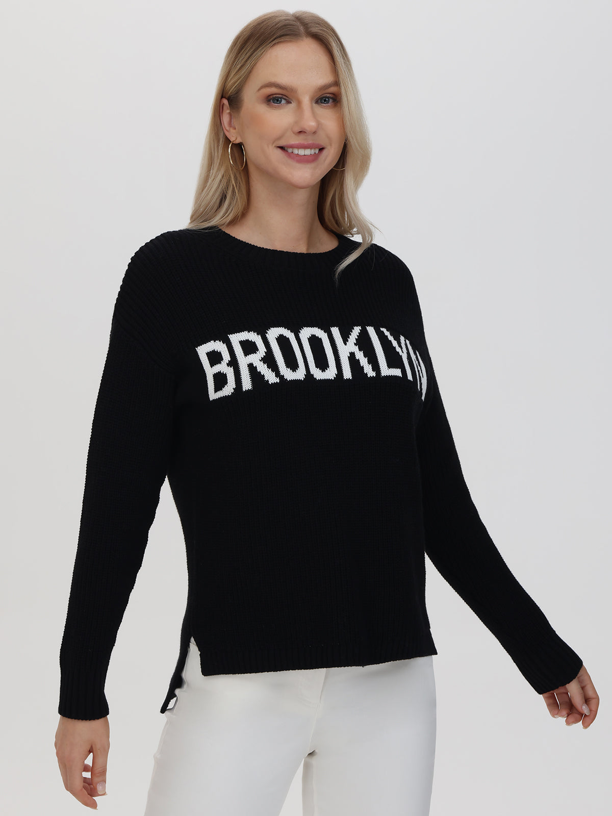 A person with long blonde hair dons a casual black 525 America sweater featuring BROOKLYN in white letters. They stylishly pair it with white pants and a smile against a plain white background.