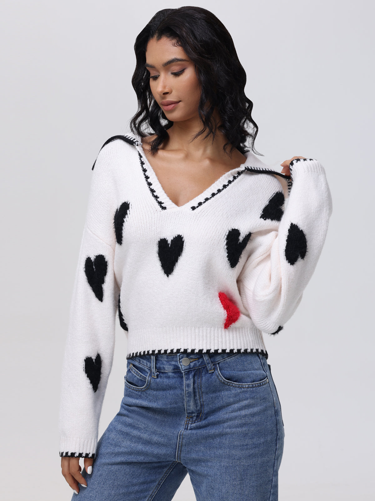 Against a neutral backdrop, someone wears a 525 America heart intarsia sweater with black hearts and a standout red one. Their long hair and blue jeans complement the V-neck design, which features black trim for balance.