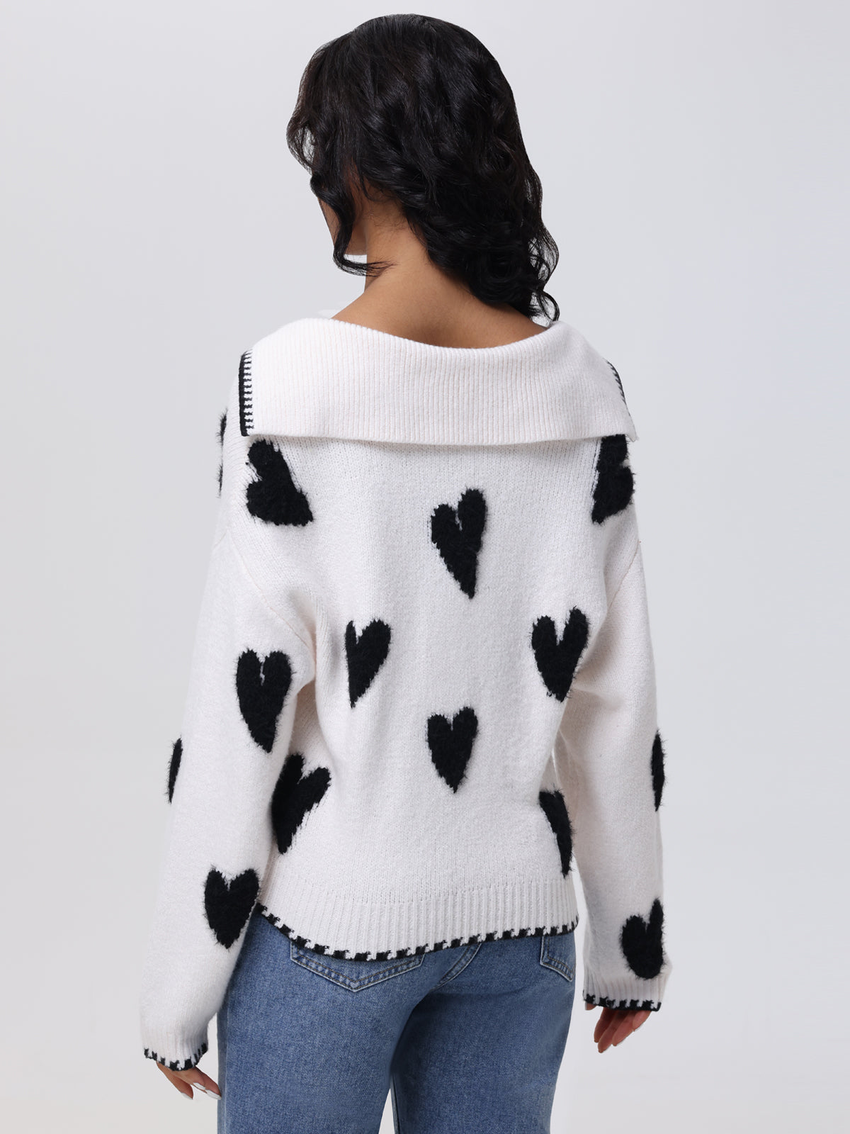 A person is seen from behind wearing a 525 America white heart intarsia sweater with an oversized nautical-style collar, ribbed cuffs, and hem. They pair it seamlessly with blue jeans against a plain white background.