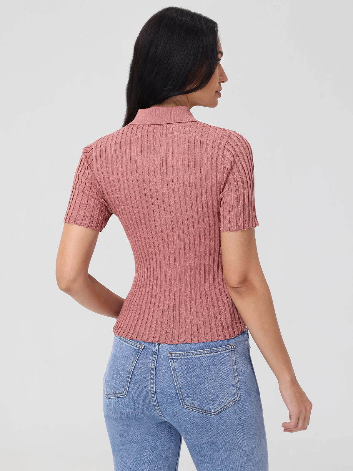 A person with long dark hair is wearing a 525 America Johnny Collar Ribbed Sweater in pink and light blue jeans, standing with their back to the camera against a plain white background, effortlessly blending style and simplicity.