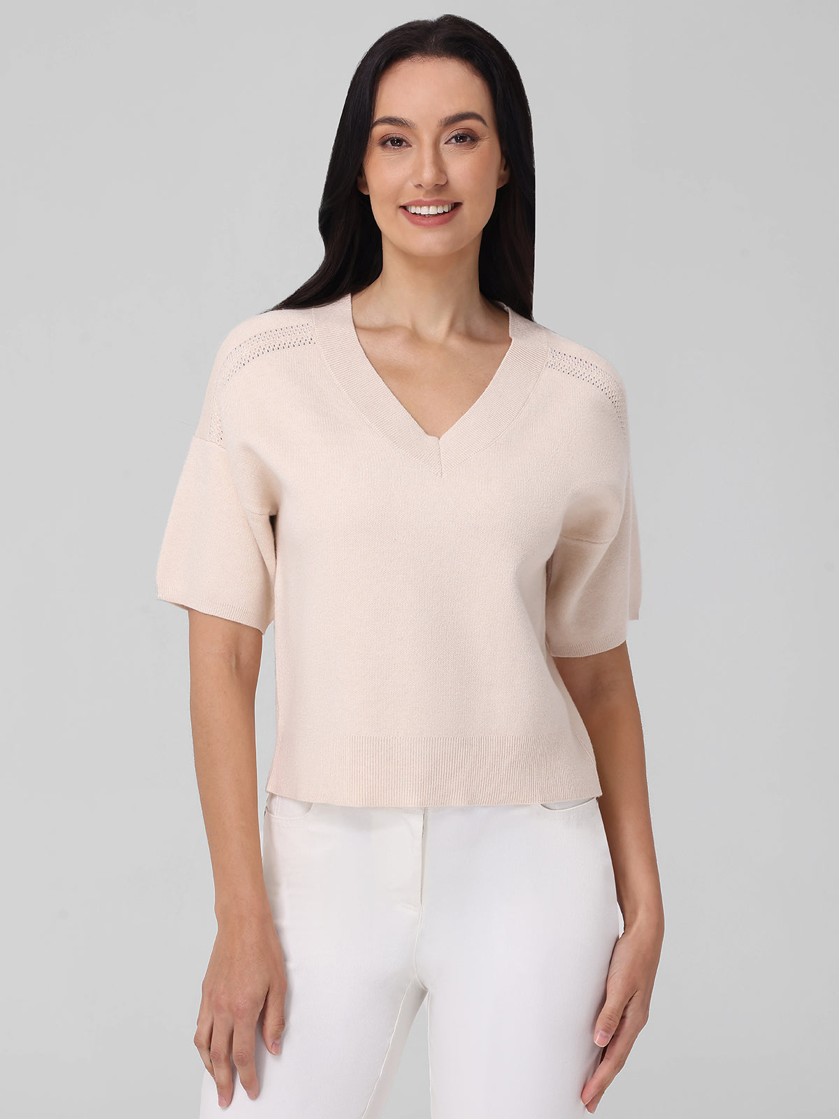 A person with long dark hair exudes timeless elegance in a light beige 525 America short-sleeved V-neck sweater, paired with white pants. They stand against a plain gray background, looking directly ahead with a slight smile.