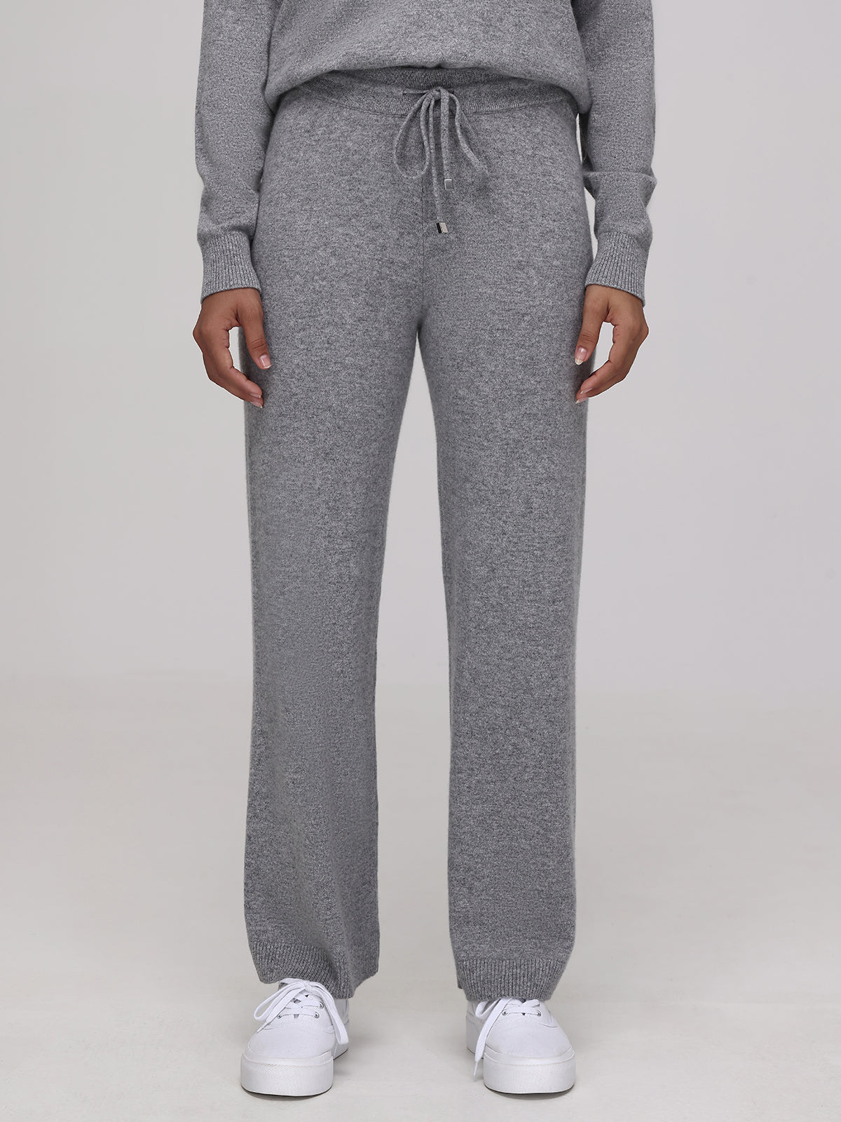 Sporting the Willow: Cashmere Straight Leg Pants in grey, complete with a drawstring waist, matched flawlessly with a coordinating top and offset by classic white sneakers. The minimalist backdrop accentuates the ensembles cozy yet luxurious fashion vibe.