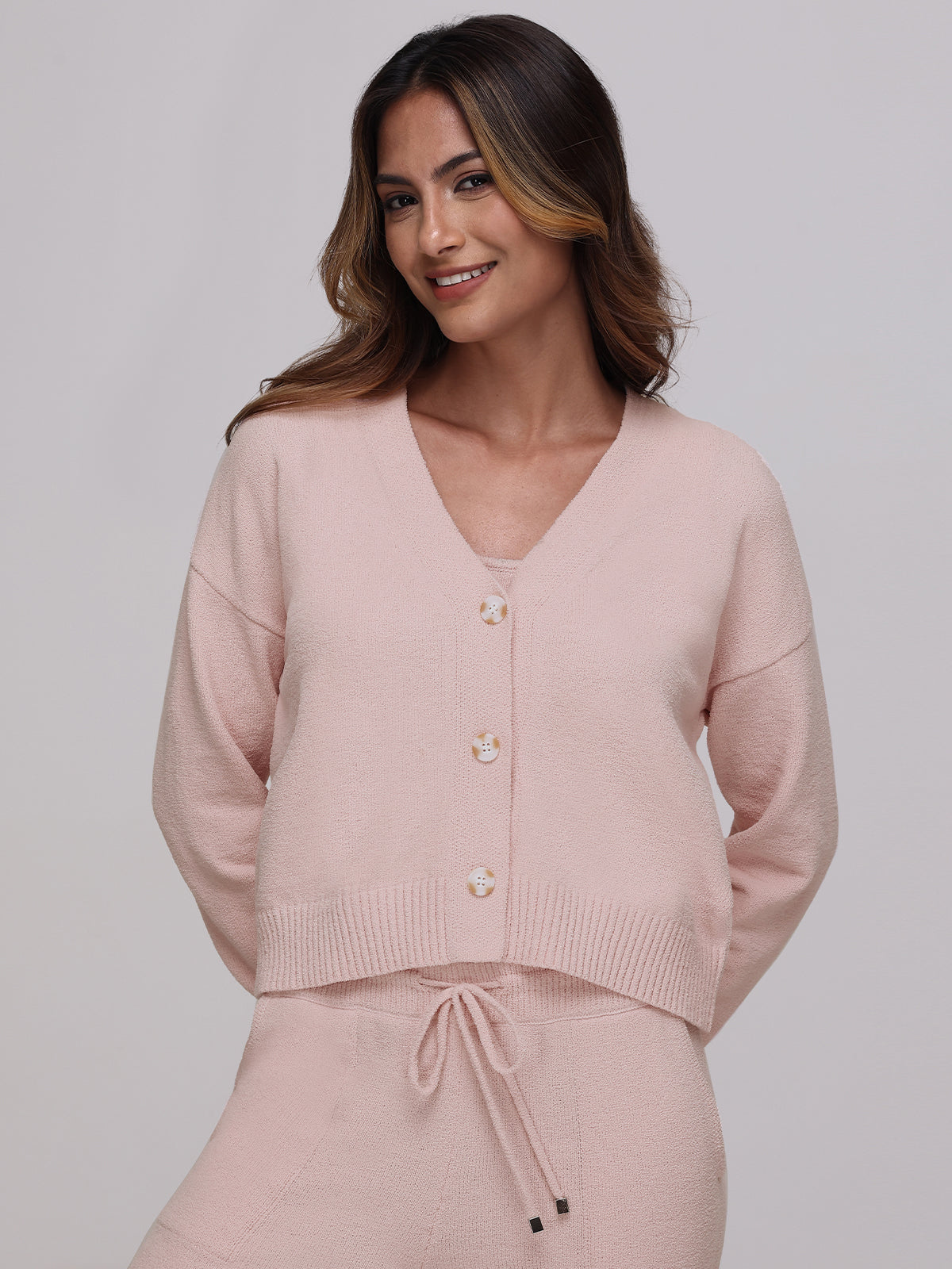 A smiling woman wears the Riley V-Neck Cardigan in light pink, paired with matching drawstring pants. Her wavy hair cascades over her shoulders against a plain backdrop—a versatile piece adding plush coziness to any wardrobe.