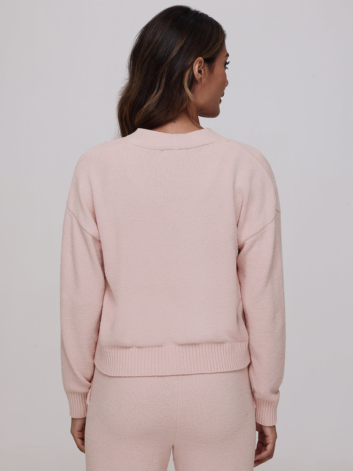 A person with long hair wears the Riley V-Neck Cardigan in light pink, radiating plush coziness with matching pants, facing away from the camera. The neutral background accentuates this versatile wardrobe staple, highlighting its texture and color.