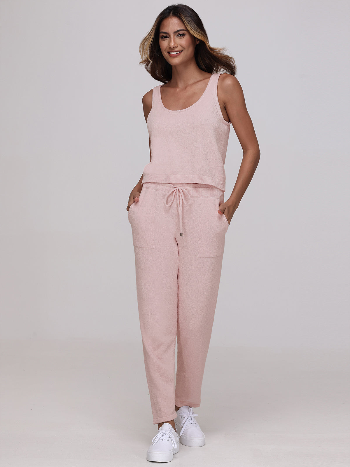 Wearing a light pink sleeveless top and matching jogger pants with drawstrings crafted from plush fabric by 525 America, a woman stands against a plain backdrop. Her hands are in her pockets as she stylishly pairs the outfit with white sneakers.