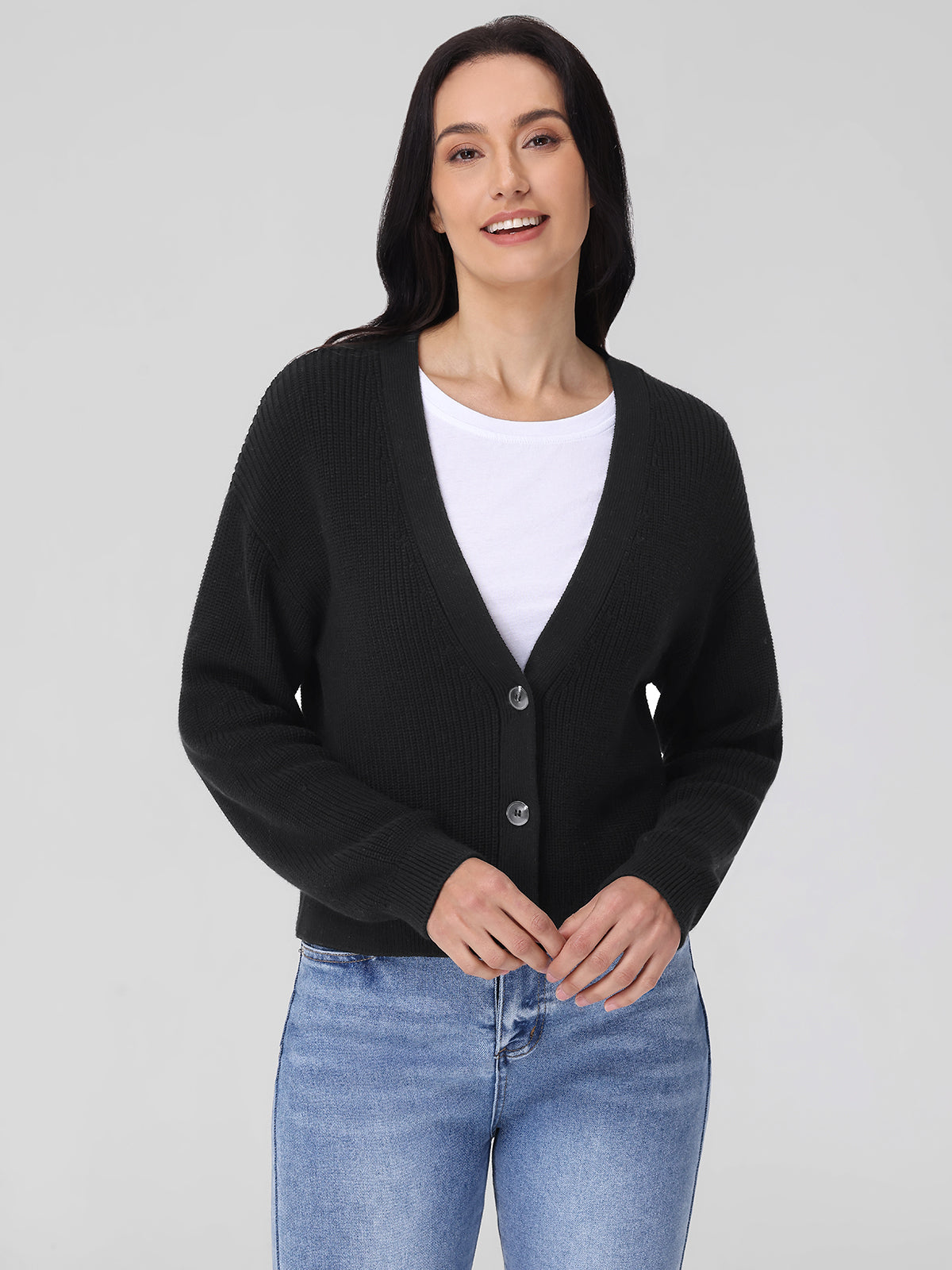 A person wears a 525 America black cardigan with shaker stitch, made from 100% cotton, over a white shirt and blue jeans against a neutral background. Their long, dark hair cascades down as they smile warmly with hands clasped in front.