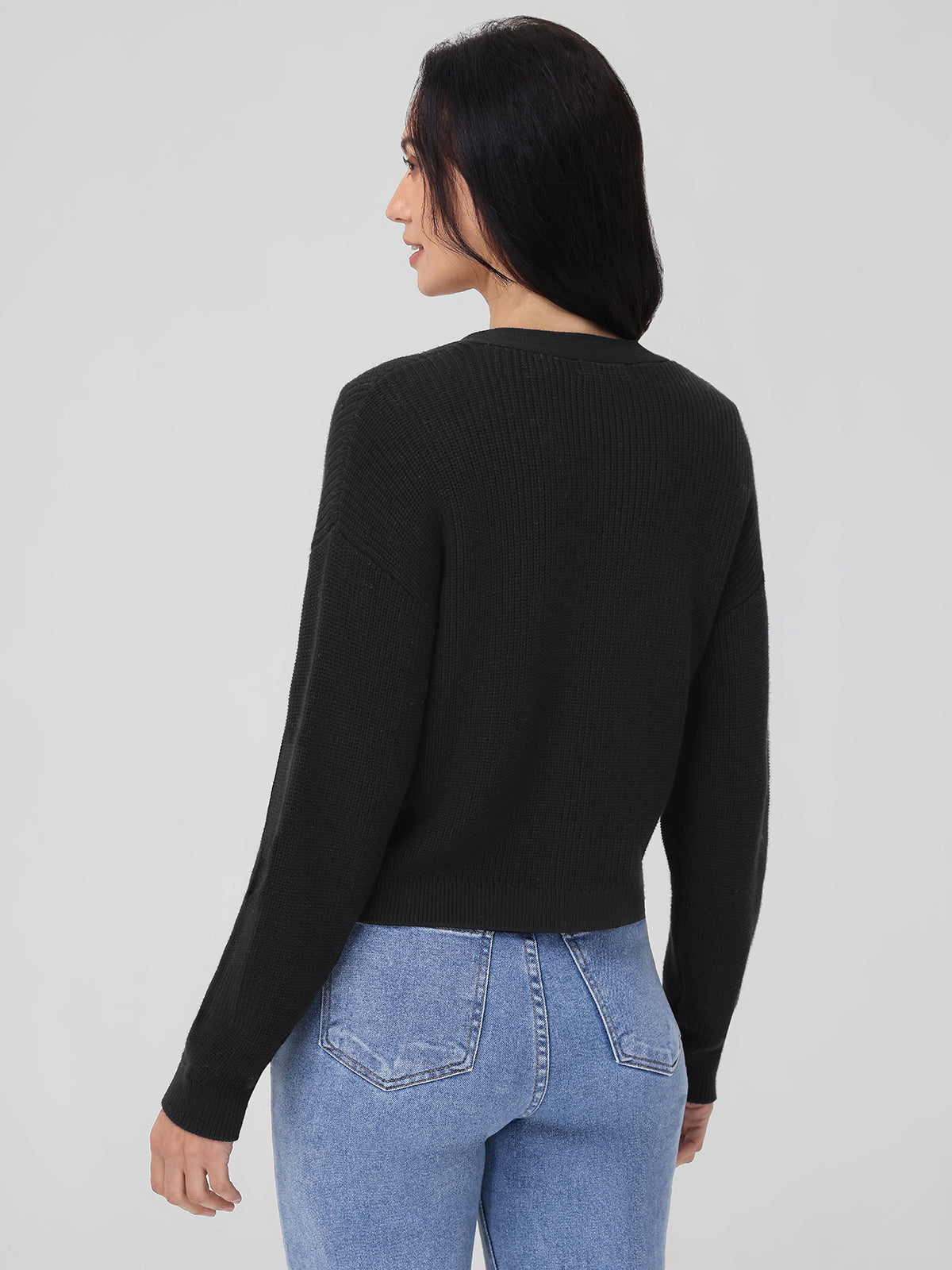 A person with long dark hair wears a black ribbed 525 America sweater made from thicker knit fabric and light blue jeans. They stand, looking over their shoulder against a plain gray backdrop.