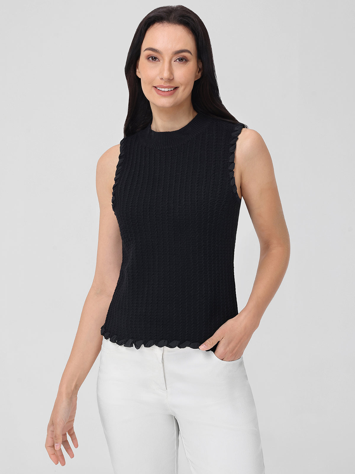 Wearing a fitted black sleeveless knit top by 525 America and white pants, a person stands against a plain background. Made from 100% cotton, the top enhances their style as they smile with one hand in their pocket.