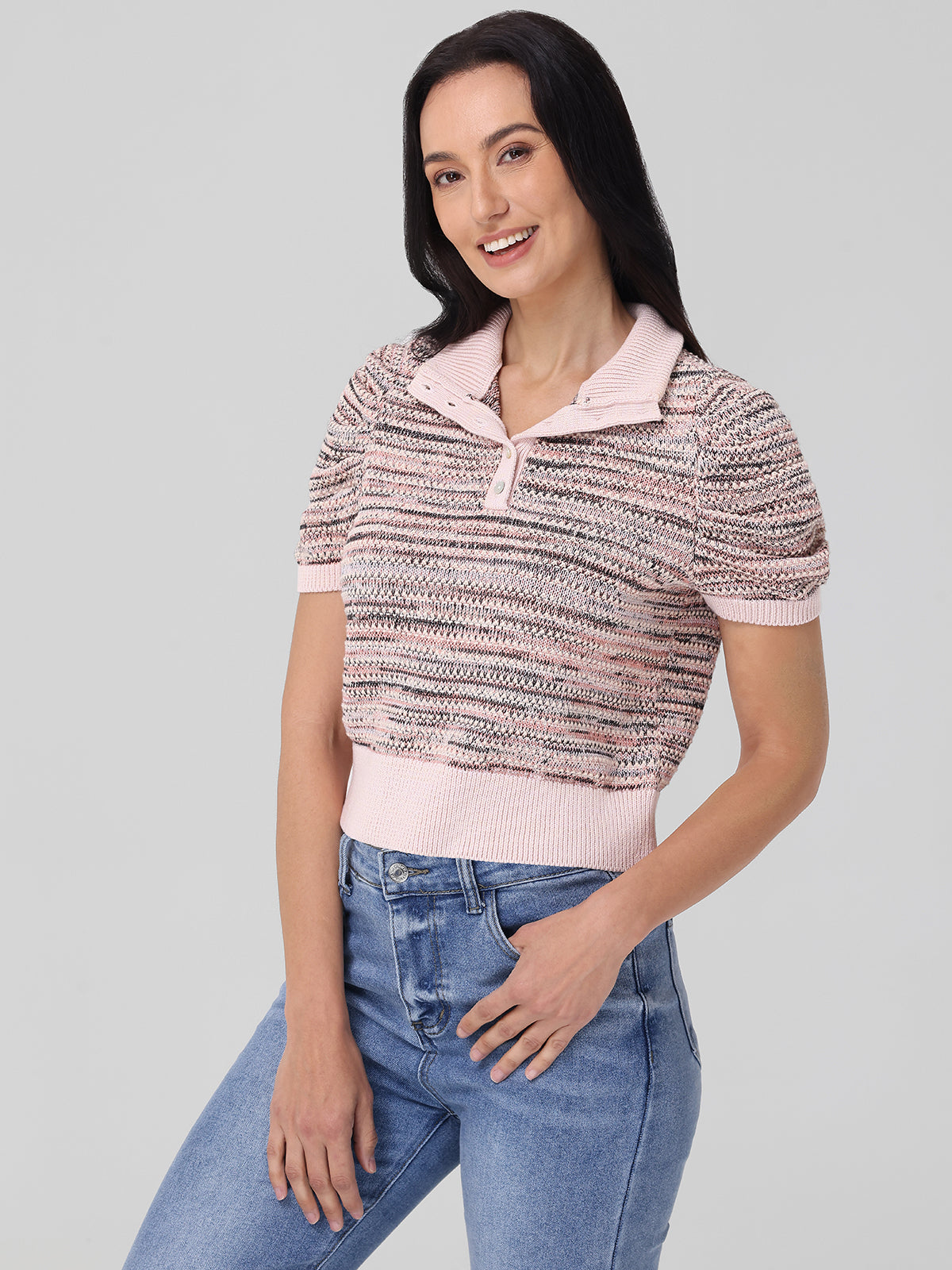 Someone with long ombre dark hair stands against a gray background, wearing a pink and gray short-sleeve 525 America sweater and blue jeans. This 100% cotton spring must-have perfectly complements their relaxed pose as they smile with one hand on their hip.