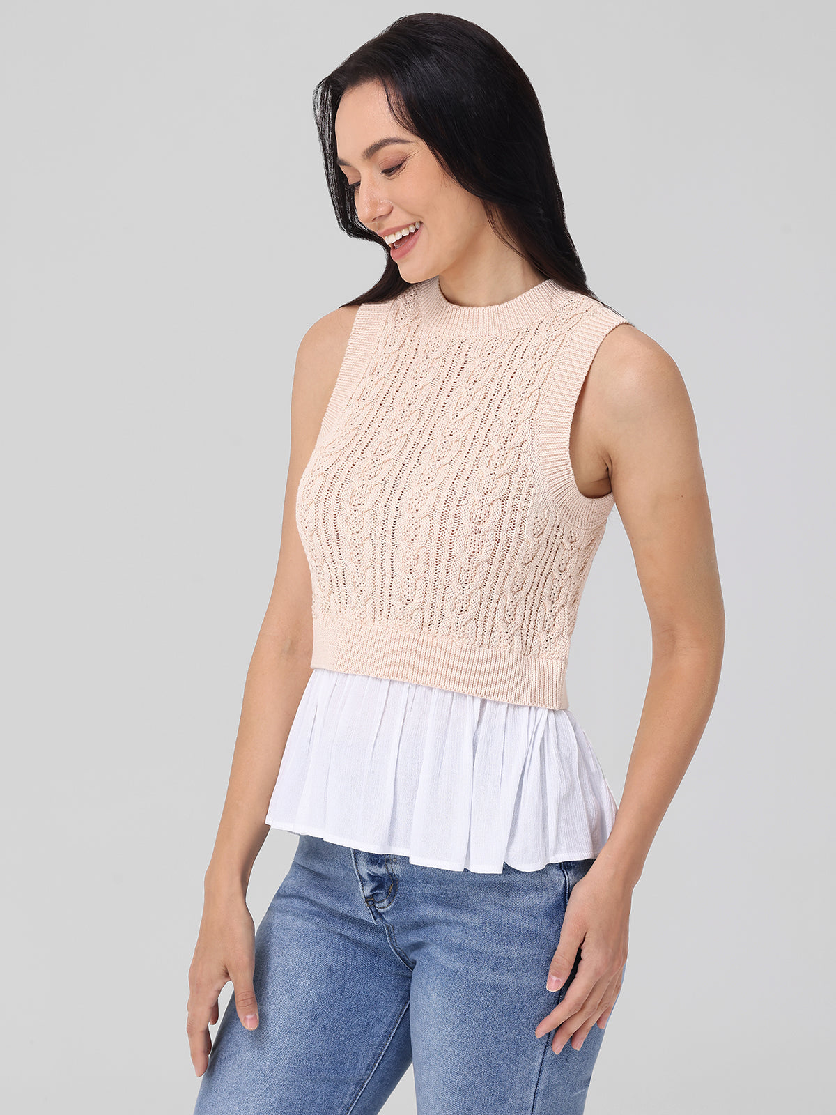 A woman in a sleeveless beige knit top by 525 America, ideal for layering, is paired with a white pleated skirt and blue jeans. She stands against a plain gray background, looking down and smiling with her hands at her sides.