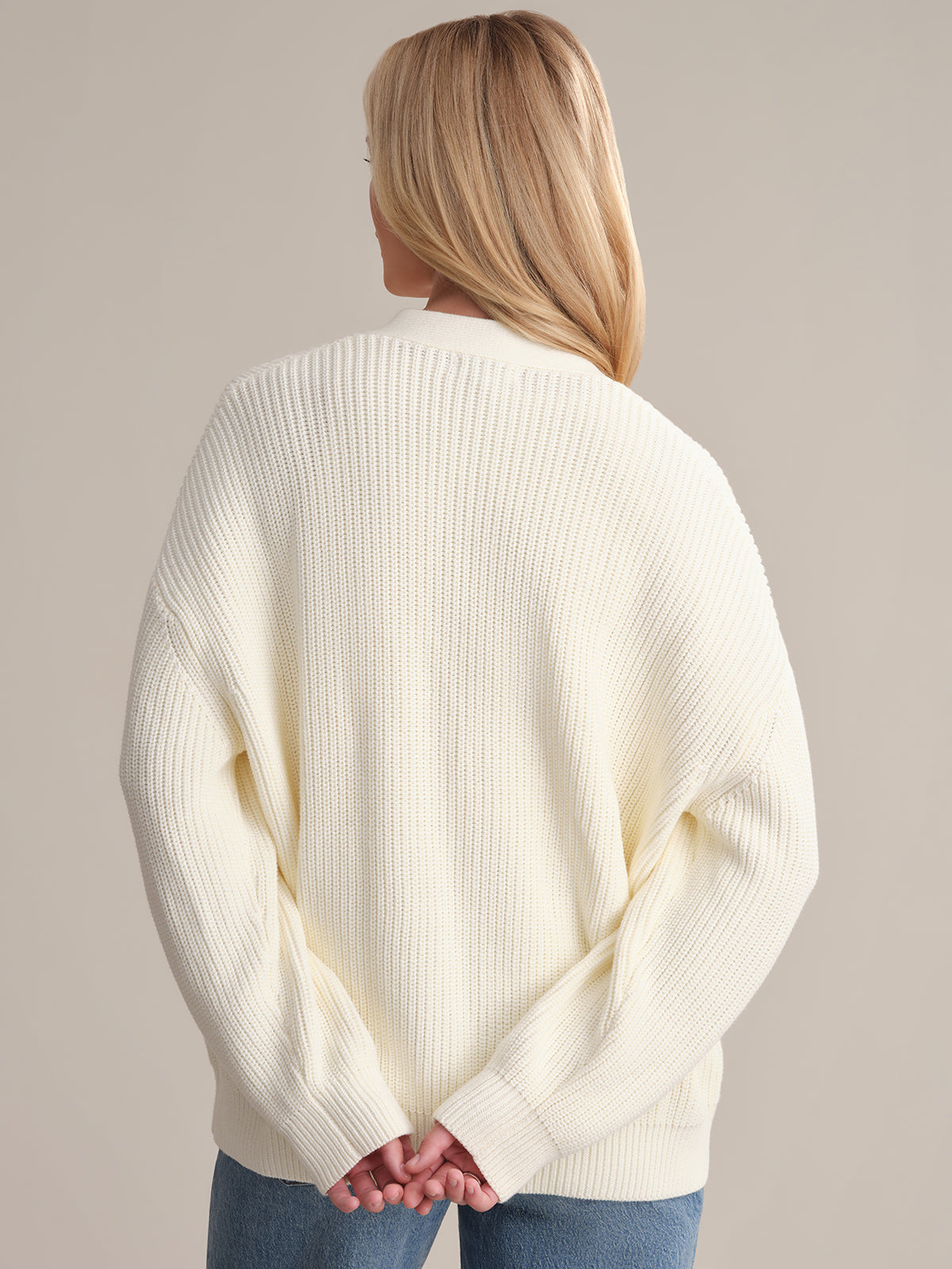 A person with long blonde hair stands facing away, wearing the cream-colored Emilia: Cotton Cocoon Cardigan in an oversized fit with blue jeans. Their hands are clasped behind, highlighting the 100% cotton texture against a plain, neutral backdrop.