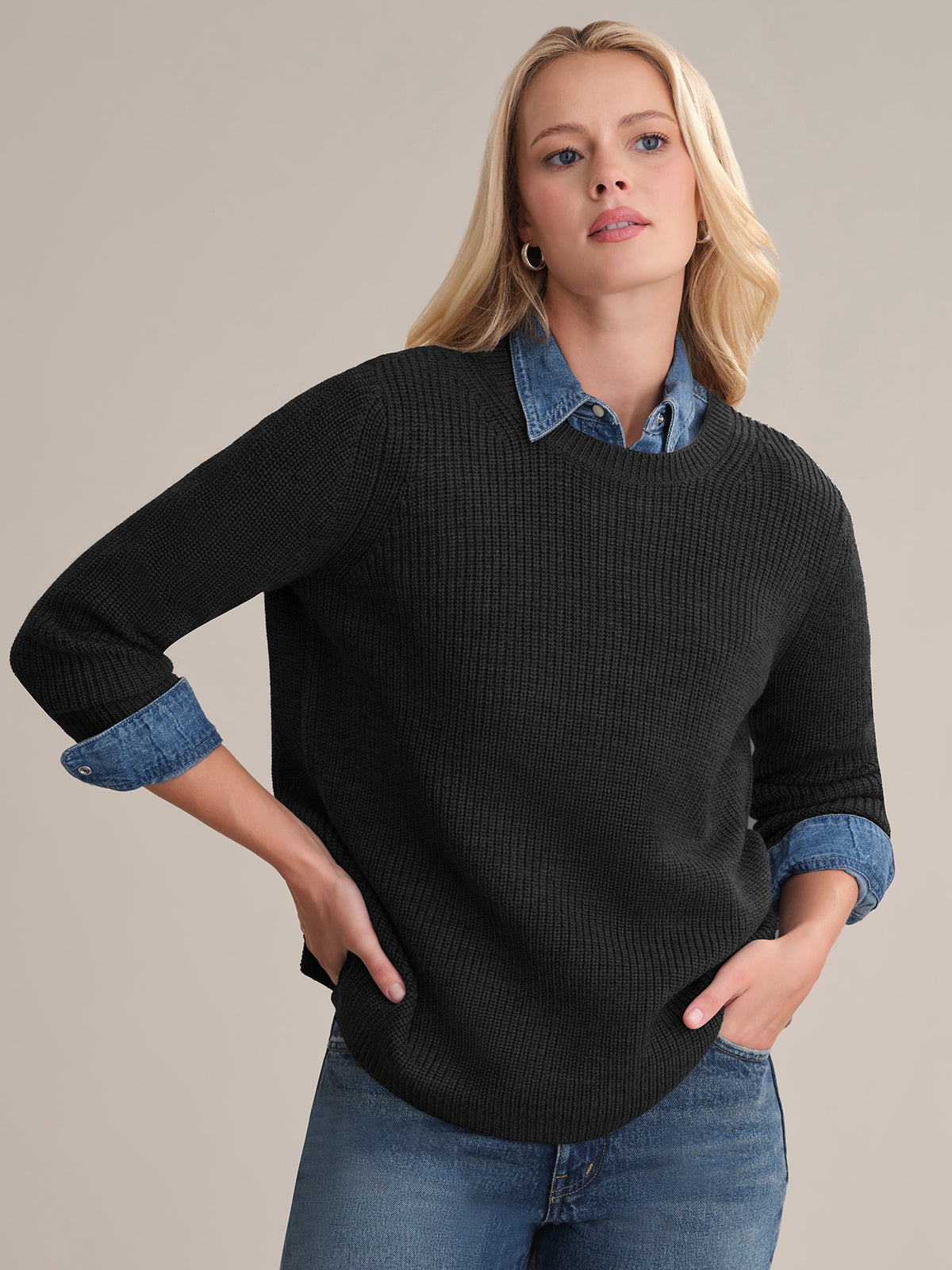 A person with long blonde hair wears the Emma: Wool Crewneck Shaker Stitch Sweater in black over a blue denim shirt and jeans. They stand against a plain background, one hand on their hip, looking slightly to the side.