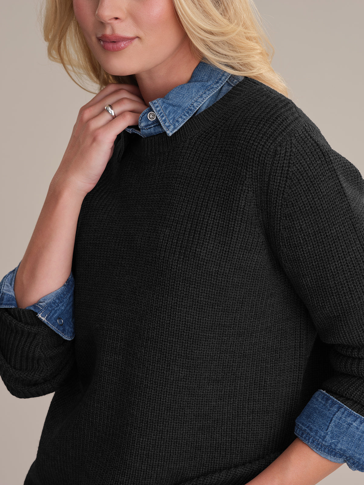 A person with long blonde hair wears the Emma: Wool Crewneck Shaker Stitch Sweater in black over a blue denim shirt. One hand touches the neckline, showcasing a silver ring. The neutral background highlights the stylish, casual layered look.