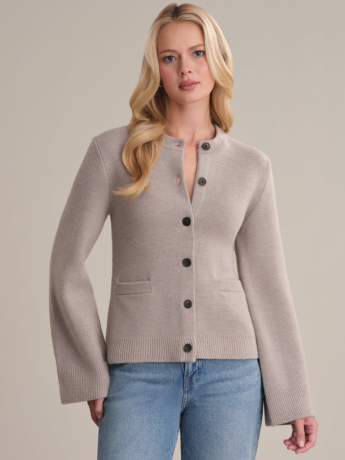 A woman with long blonde hair stands against a plain background, wearing the Catherine Wool Cardigan featuring black buttons, two small front pockets, a round neckline, and blue jeans. This sophisticated cardigan adds a timeless touch to her look.