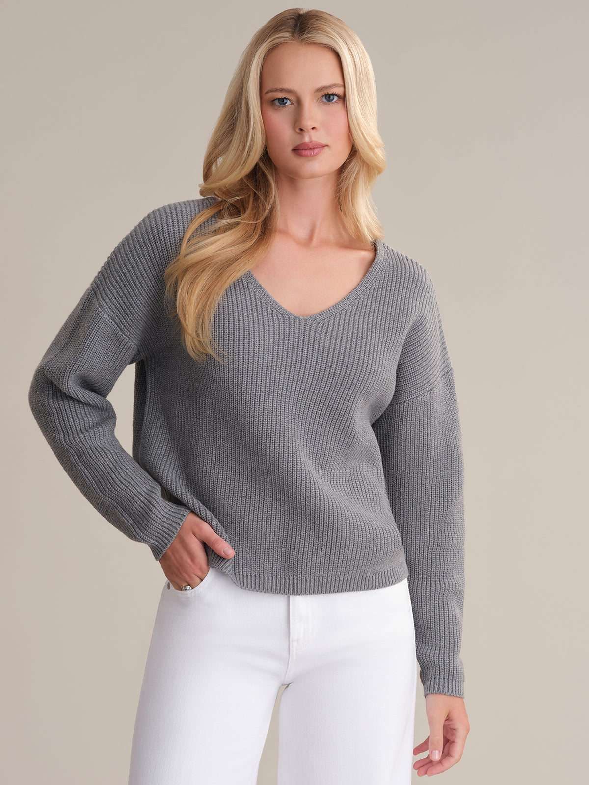 A person with long blonde hair wears a 525 America Ava V-Neck Sweater in gray ribbed shaker stitch and white pants, standing against a neutral background, hand in pocket and relaxed expression.