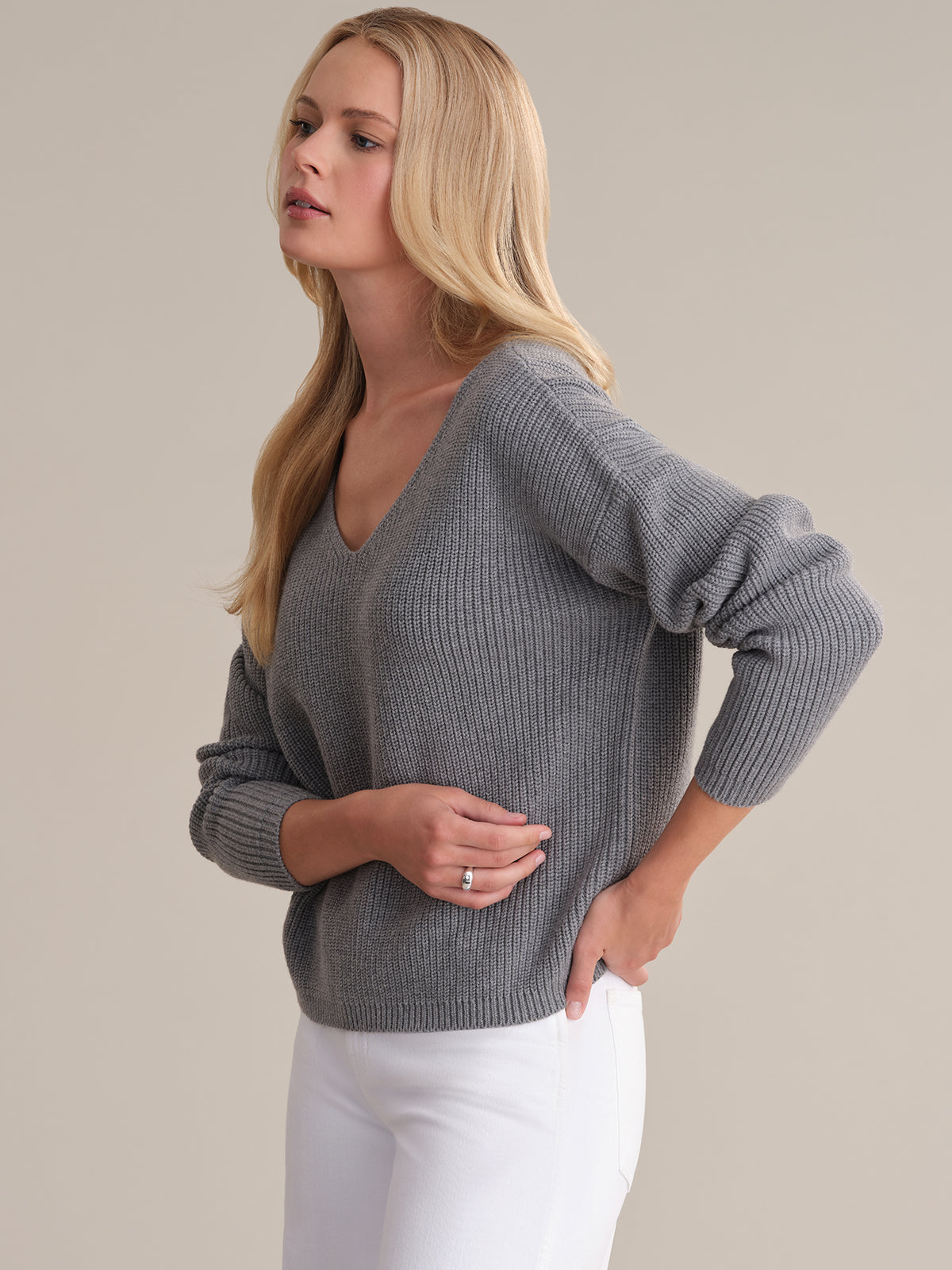 A person with long blonde hair wears a 525 Americas Ava V-Neck Sweater in grey ribbed shaker stitch design, paired with white pants. They pose against a plain background, hand on hip, gazing slightly to the side.