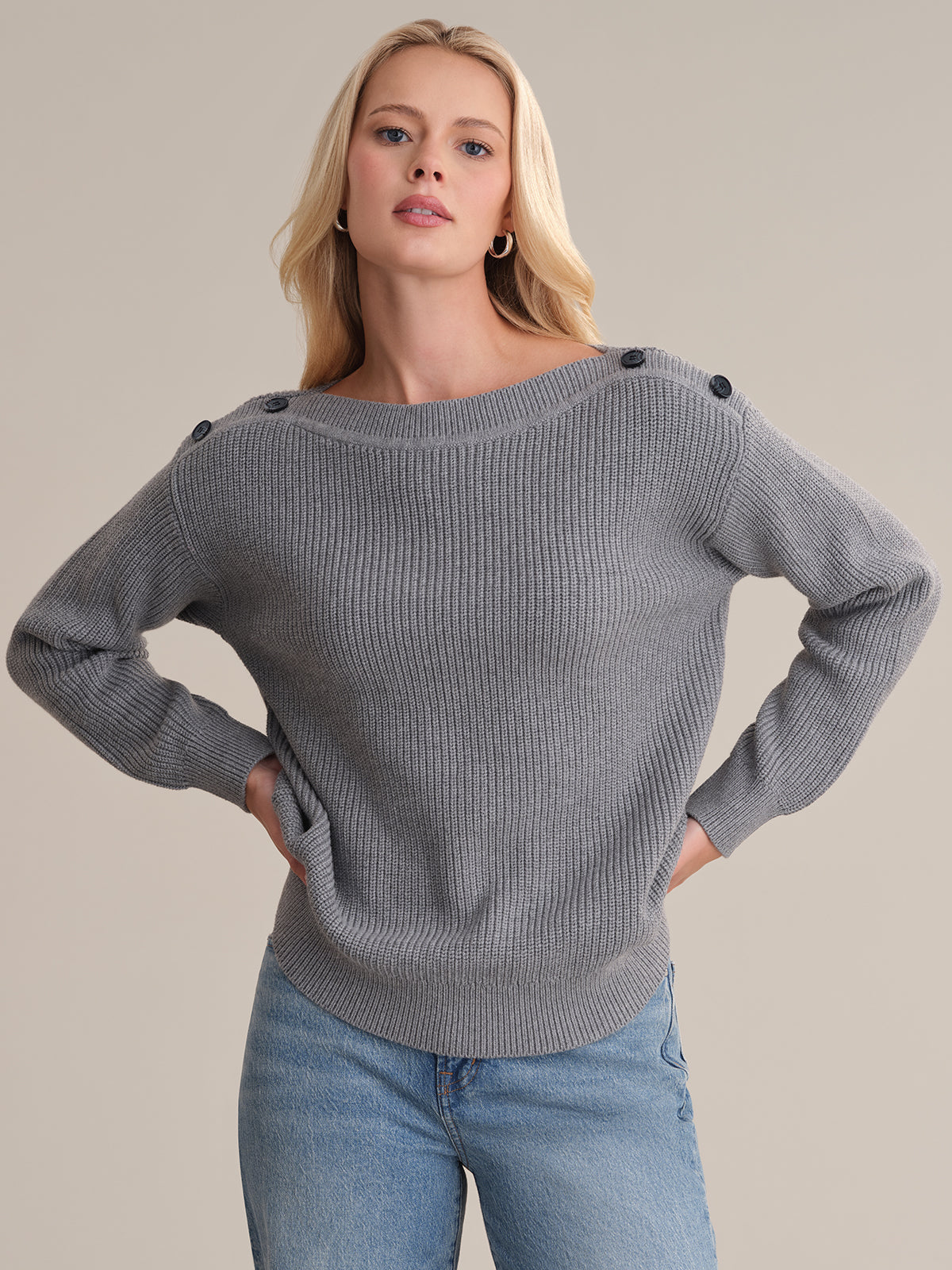 A person with long blonde hair stands against a plain background, wearing a 525 America versatile gray shaker stitch sweater featuring button details on the shoulders and light blue jeans. They look directly at the viewer with hands in pockets.