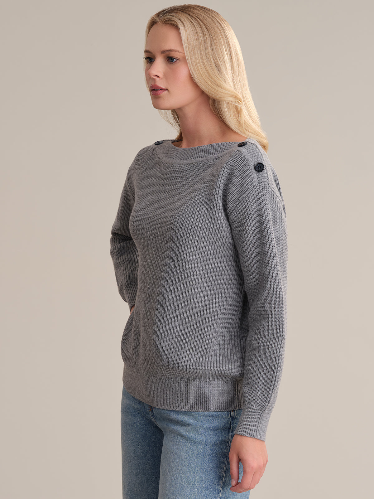 A person with long blond hair is wearing a versatile gray ribbed sweater by 525 America, featuring button details on the shoulders, paired with blue jeans. They are standing against a plain beige background, facing slightly to the right.