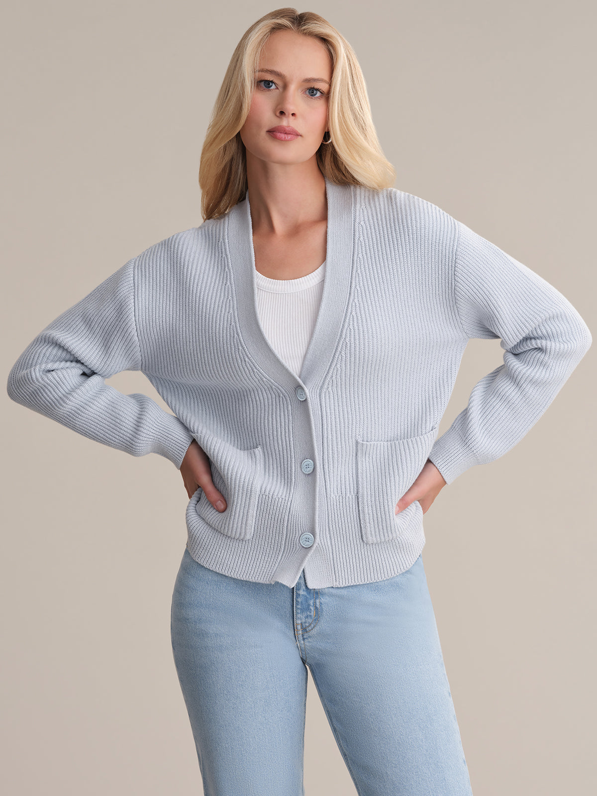 A person with long blonde hair wears a light blue v-neck cardigan by 525 America over a white top and matching jeans, posing with hands on hips against a plain beige background.