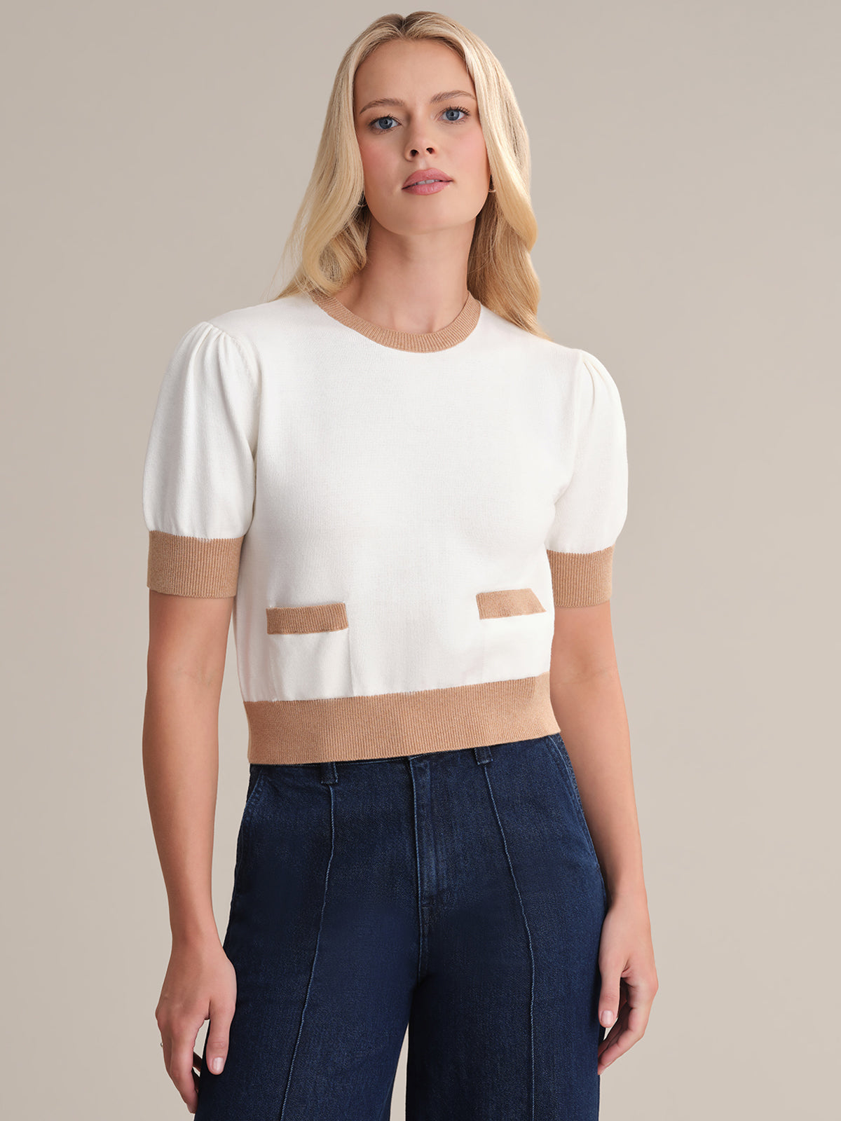 A woman with long blonde hair models a feminine 525 America pullover, a white short-sleeved sweater with tan cuffs and hem, showcasing two rectangular pockets. Made from 100% cotton, she pairs it stylishly with dark blue high-waisted jeans against a plain beige backdrop.