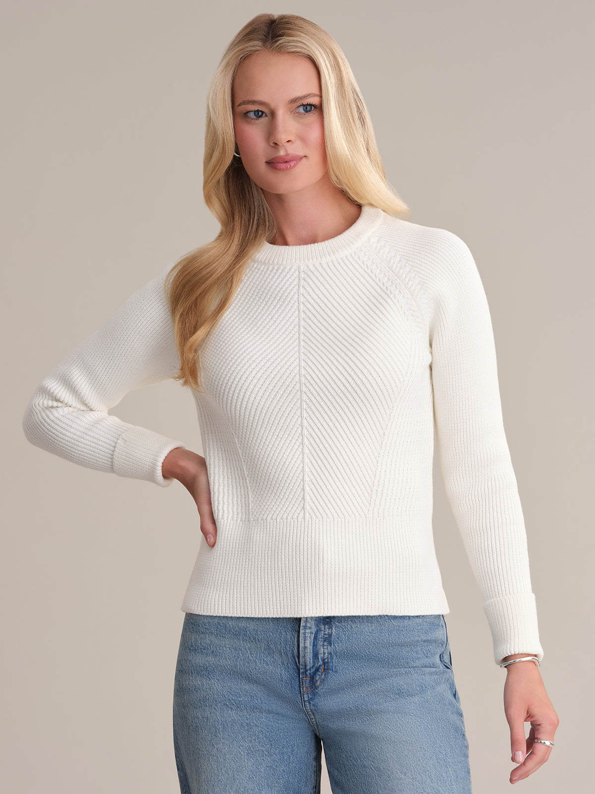 A person with long, blonde hair is wearing a sophisticated white chevron rib texture sweater by 525 America paired with blue jeans. They have one hand on their hip and are looking off to the side against a plain beige background.