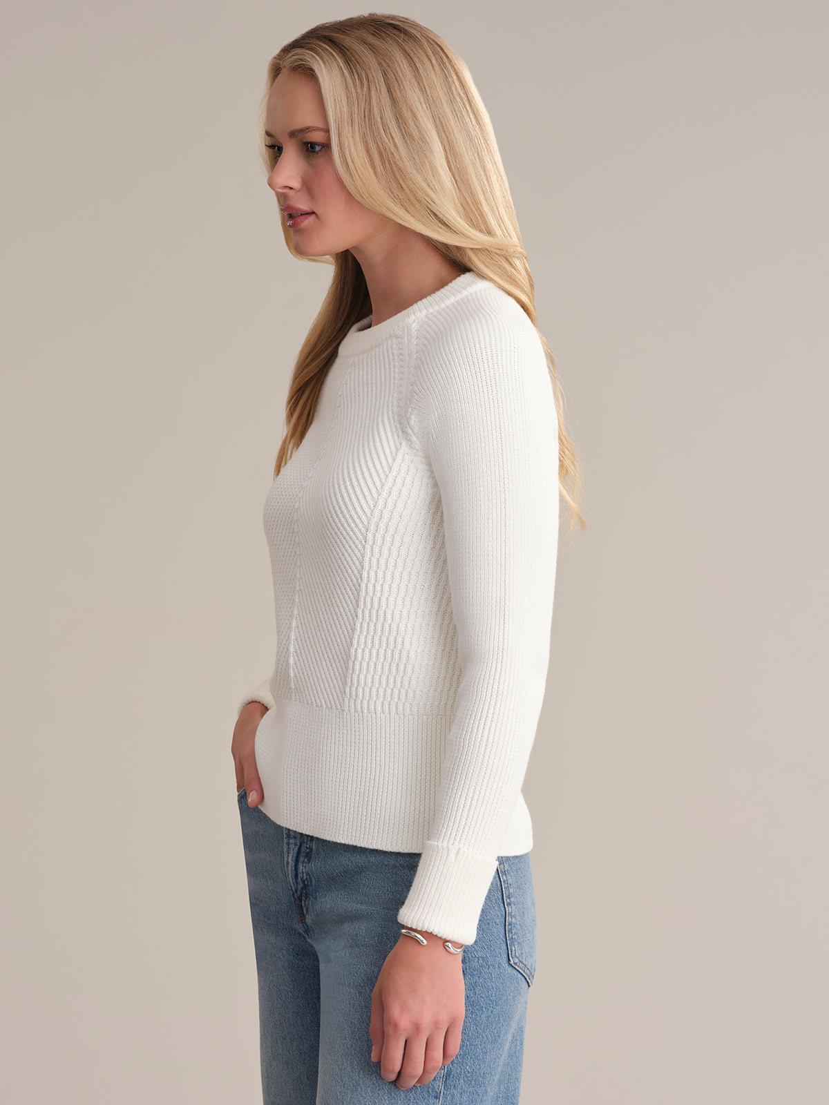 A person with long blonde hair, facing left, wears a sophisticated chevron rib texture sweater from 525 America and classic-fit blue jeans, their hands in the pockets. The background is a plain, light neutral color.