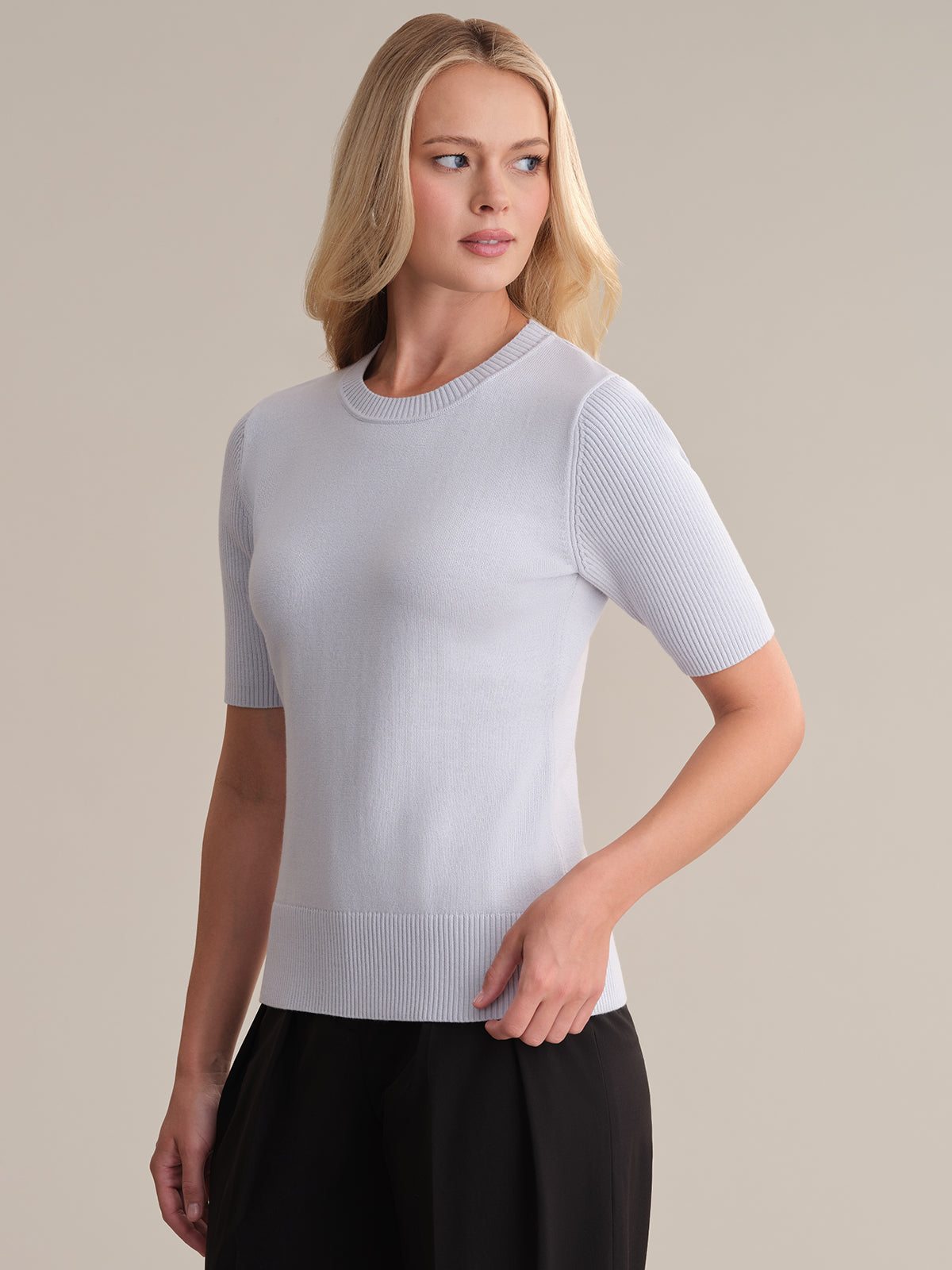 A person with long blonde hair wears a light gray short-sleeve cotton sweater from 525 America, highlighting a fitted silhouette, paired with black pants. They stand against a plain beige background, looking slightly to the side.