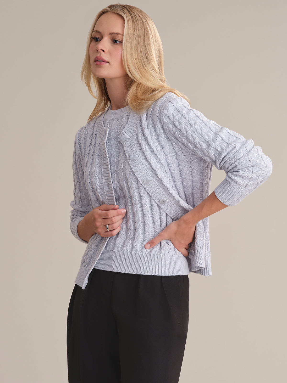 A person with long blonde hair is wearing a 525 America light blue cotton cable-knit sweater over a matching top and black pants, standing against a neutral background with one hand on their hip.