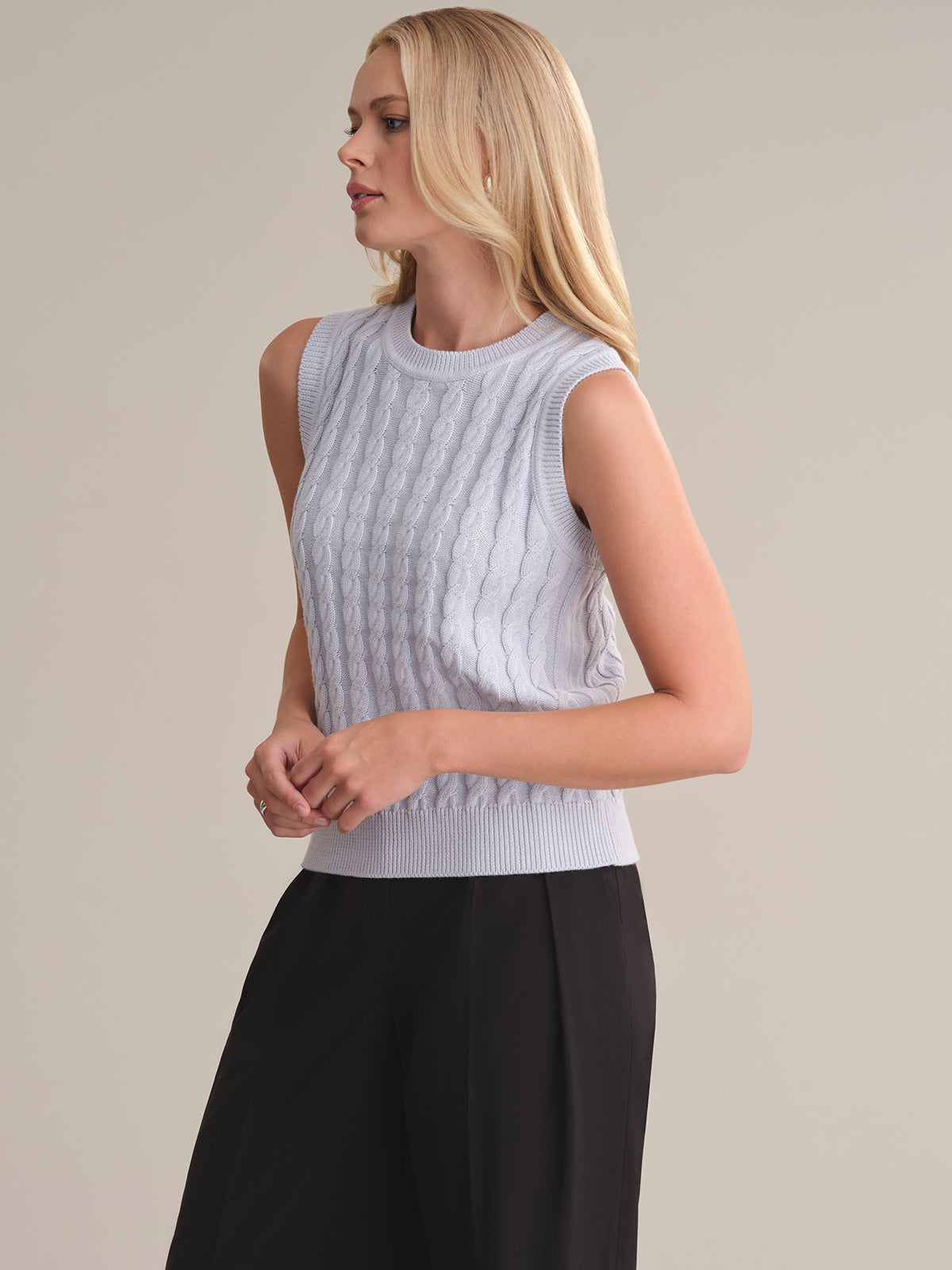 Someone with long blonde hair is modeling a sleeveless, light blue cable-knit sweater in a relaxed silhouette by 525 America paired with black pants, standing in a neutral pose against a plain background.