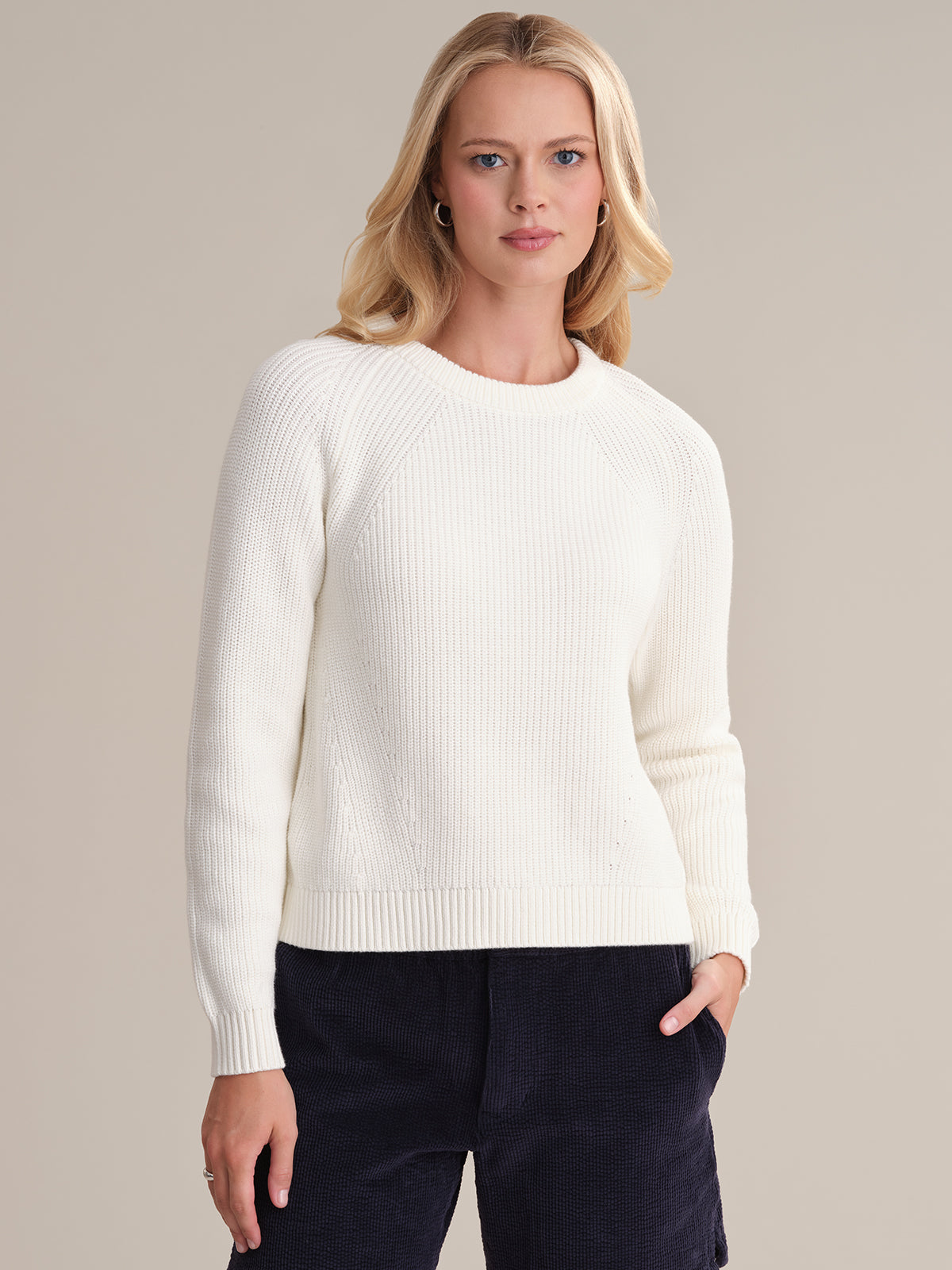 Someone with long blonde hair wears a 525 America white cotton shaker stitch sweater with raglan sleeves and dark pants. With a hand in their pocket, they pose against a neutral background, showcasing the fitted silhouette of their outfit.