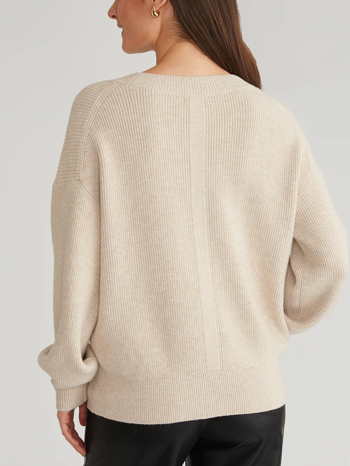 A person is seen from behind wearing a beige, ribbed, long-sleeved sweater with a relaxed fit and back seam by 525 America. Paired with black pants, their shoulder-length brown hair enhances the effortless elegance of their look.