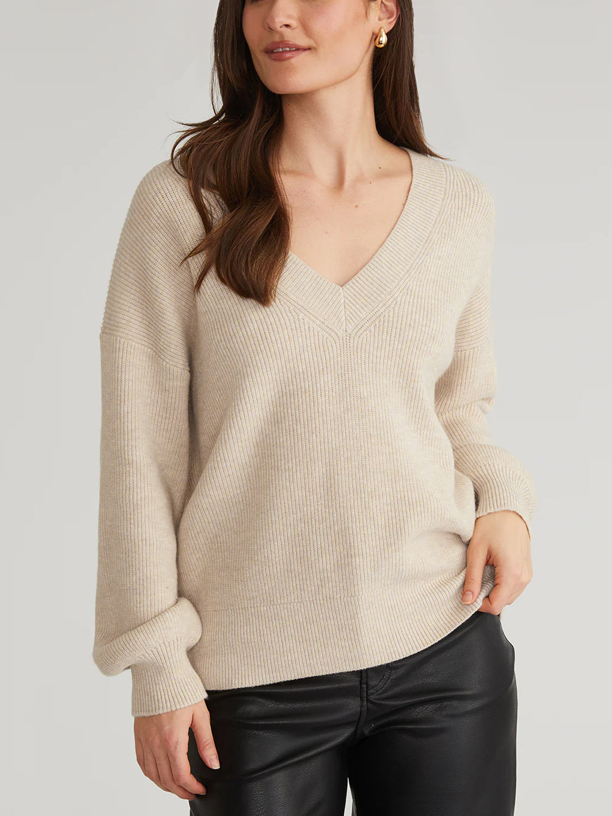 A woman confidently poses against a neutral backdrop wearing a relaxed-fit beige V-neck sweater by 525 America and black pants, her hands on her hips. Her long hair cascades down, complementing the earring in her visible ear.