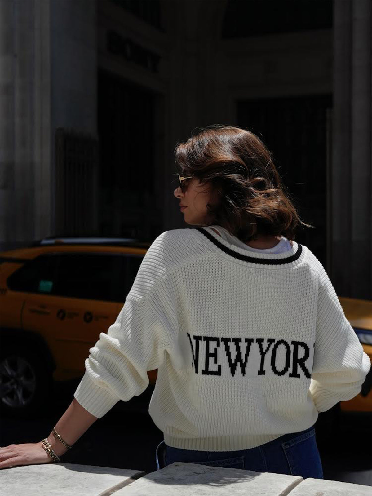 Facing away, a person with shoulder-length hair and sunglasses wears a relaxed fit 100% cotton sweater by 525 America, featuring NEW YORK on the back. Behind them, a yellow taxi approaches a building entrance, highlighting the citys vibrant energy.