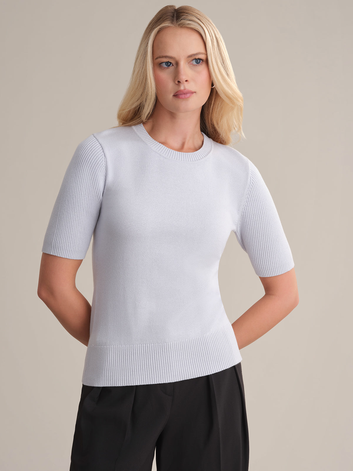 A person with long blonde hair is wearing a light gray, short-sleeve 100% cotton sweater from 525 America. It features a fitted silhouette and is paired with black pants. They stand against a plain background, hands behind their back, gazing slightly to the side.