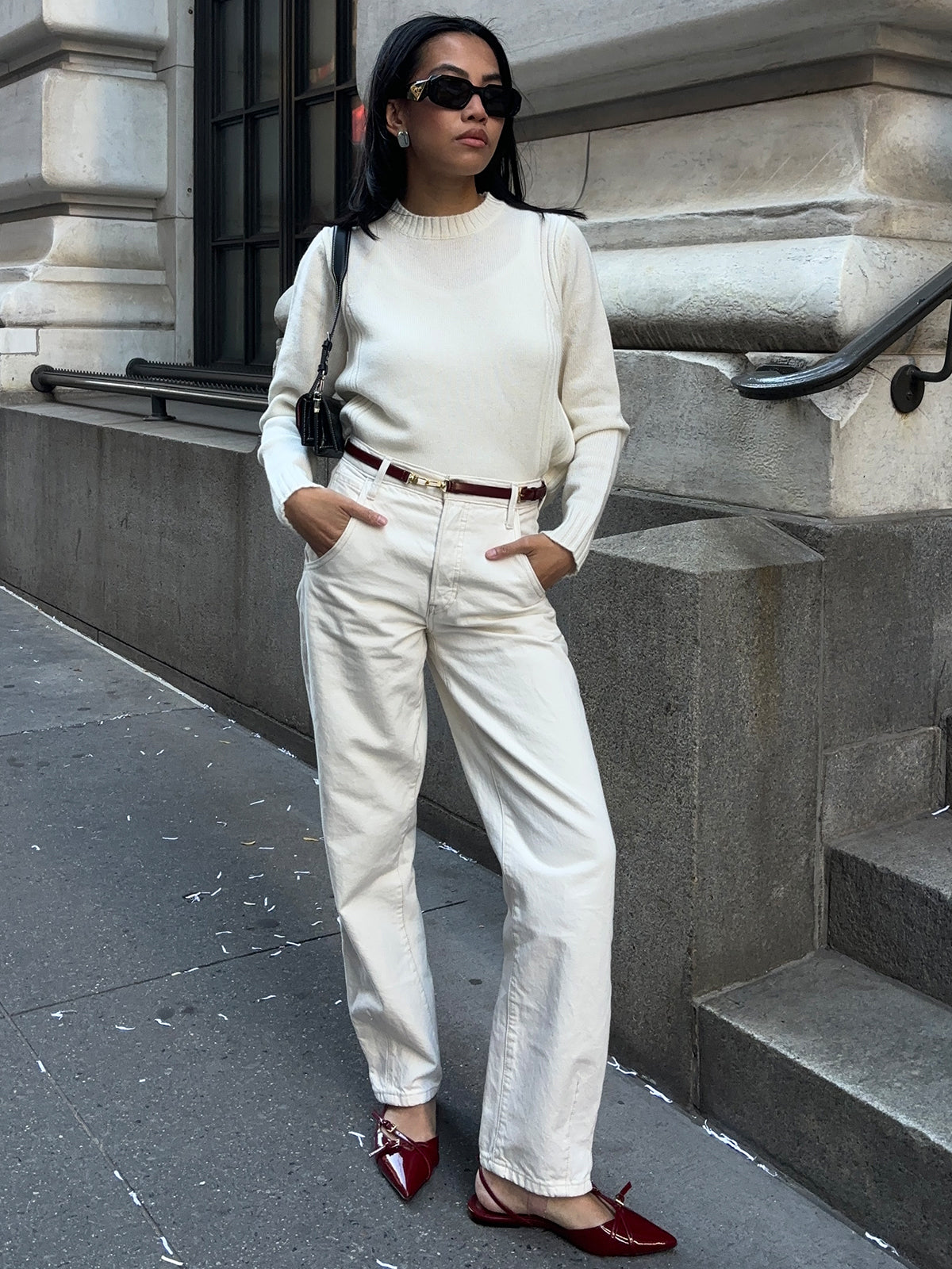 Outdoors on a city sidewalk, someone is styled in a chic all-white outfit from 525 America, showcasing a luxurious cashmere crewneck sweater and high-waisted pants. Red pointed shoes, dark sunglasses, and a crossbody bag complete the look as they casually rest their hands in pockets.