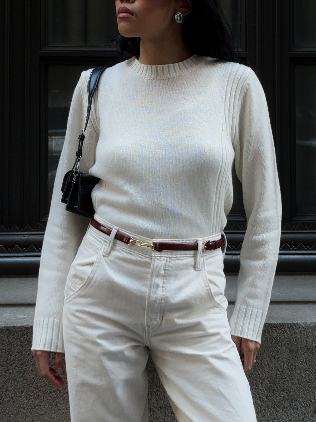 Wearing a cream cashmere crewneck sweater from 525 America and white jeans, a person stands by a dark window. They accessorize with a small black shoulder bag and burgundy belt against the backdrop of stone ledges and ornate window trim.