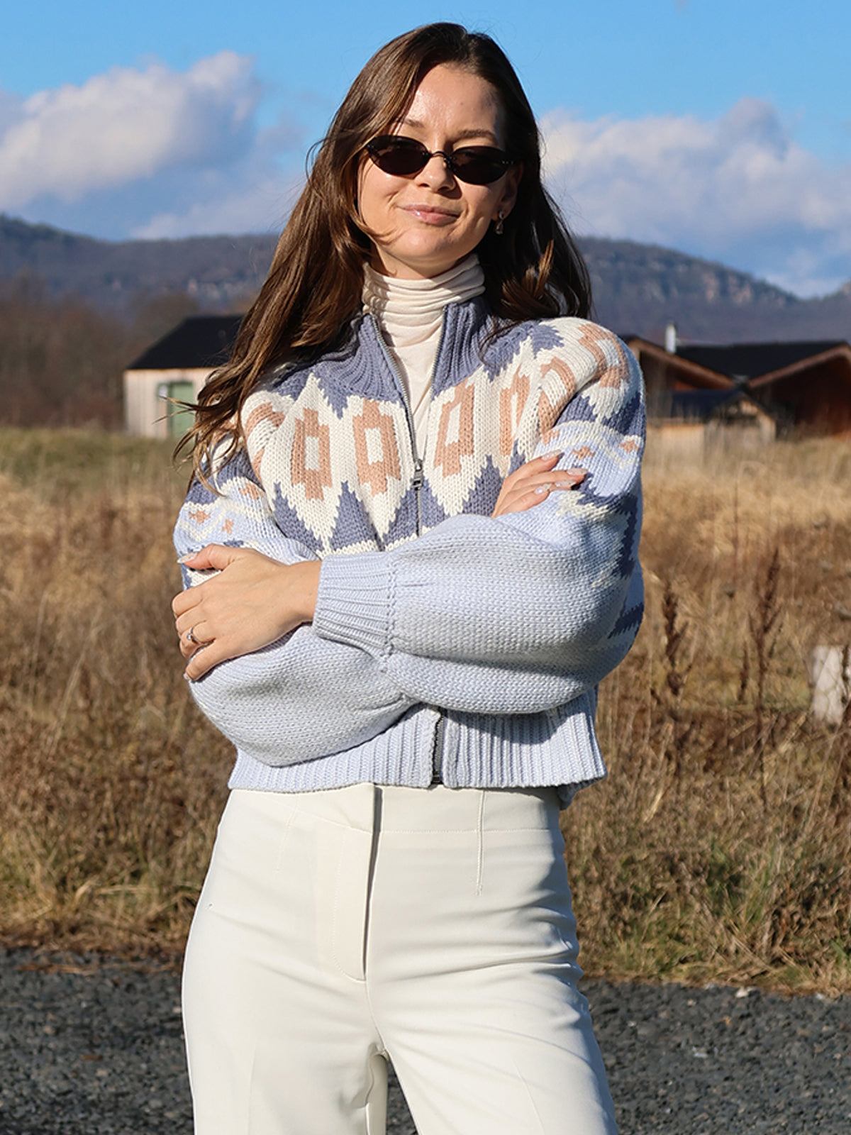 In a Victoria: Fair Isle Sweater Bomber Jacket, 100% acrylic and adorned with winter patterns, a person stands confidently outdoors with crossed arms and sunglasses, set against a backdrop of hills, houses, and a partly cloudy rural sky.