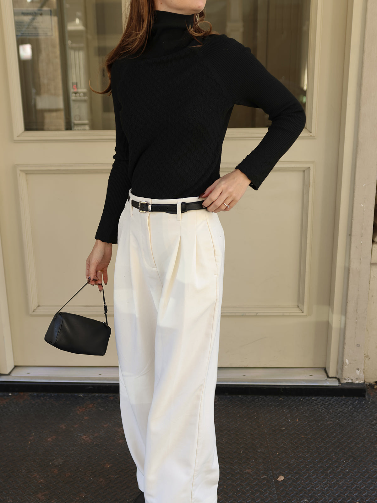 A person with long hair stands before a building with beige doors, wearing a soft knit black turtleneck sweater by 525 America and white pleated trousers. They carry a small black handbag and have one hand on their hip.