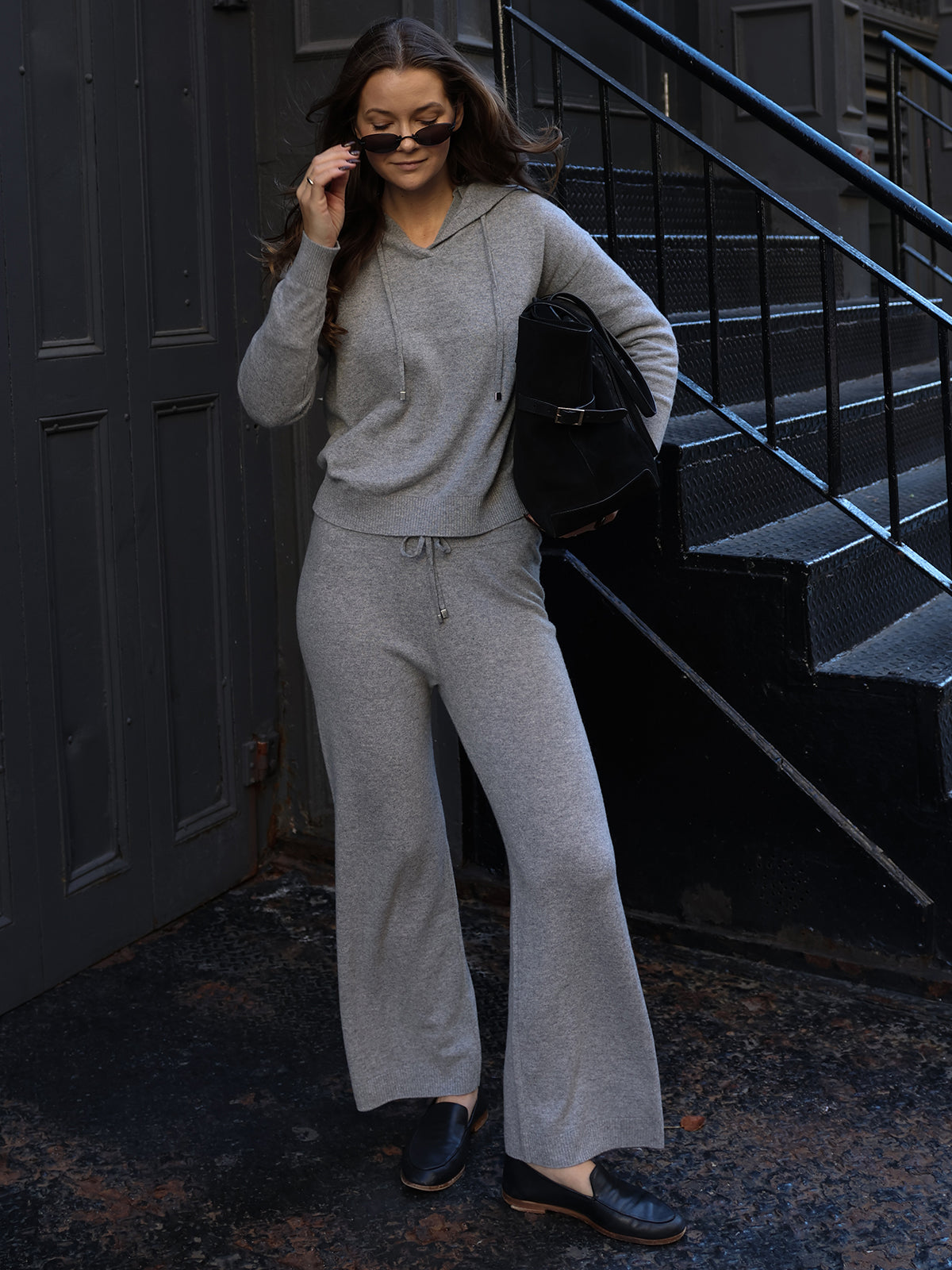 A person in a gray hoodie and Willow: Cashmere Straight Leg Pants stands on a textured sidewalk with sunglasses and a black bag. They epitomize luxury fashion with a staircase in the background.